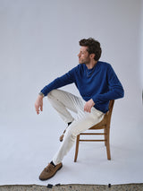 A man wearing a blue long-sleeve shirt and BGvinci Bianco Jeans - White by Blue de Gênes sits sideways on a wooden chair against a plain white backdrop, looking to his left. He pairs his outfit with brown shoes for all-day comfort.