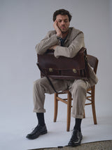 A man wearing the Blue de Gênes BGjazzy Ola Jacket in Dark Sand sits on a wooden chair, resting his head on one hand with a bored expression. He holds a large brown briefcase and wears black shoes. The background is plain and light-colored.