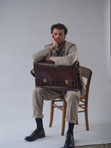 A man in a BGjazzy Ola Jacket - Dark Sand by Blue de Gênes sits on a wooden chair, head resting on one hand, holding a large leather briefcase on his lap, looking tired or bored against a plain background.