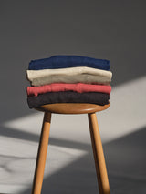 A stack of four BGmonty Knit crewneck jumpers by Blue de Gênes in coral, navy blue, beige, and black sits on a wooden stool as soft natural light casts subtle shadows in the background.