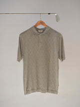 The BGwally Diamond Polo in Shadow Green by Blue de Gênes, featuring a subtle diamond-knit pattern, is displayed on a wooden hanger suspended from a metal golf club against a plain background.