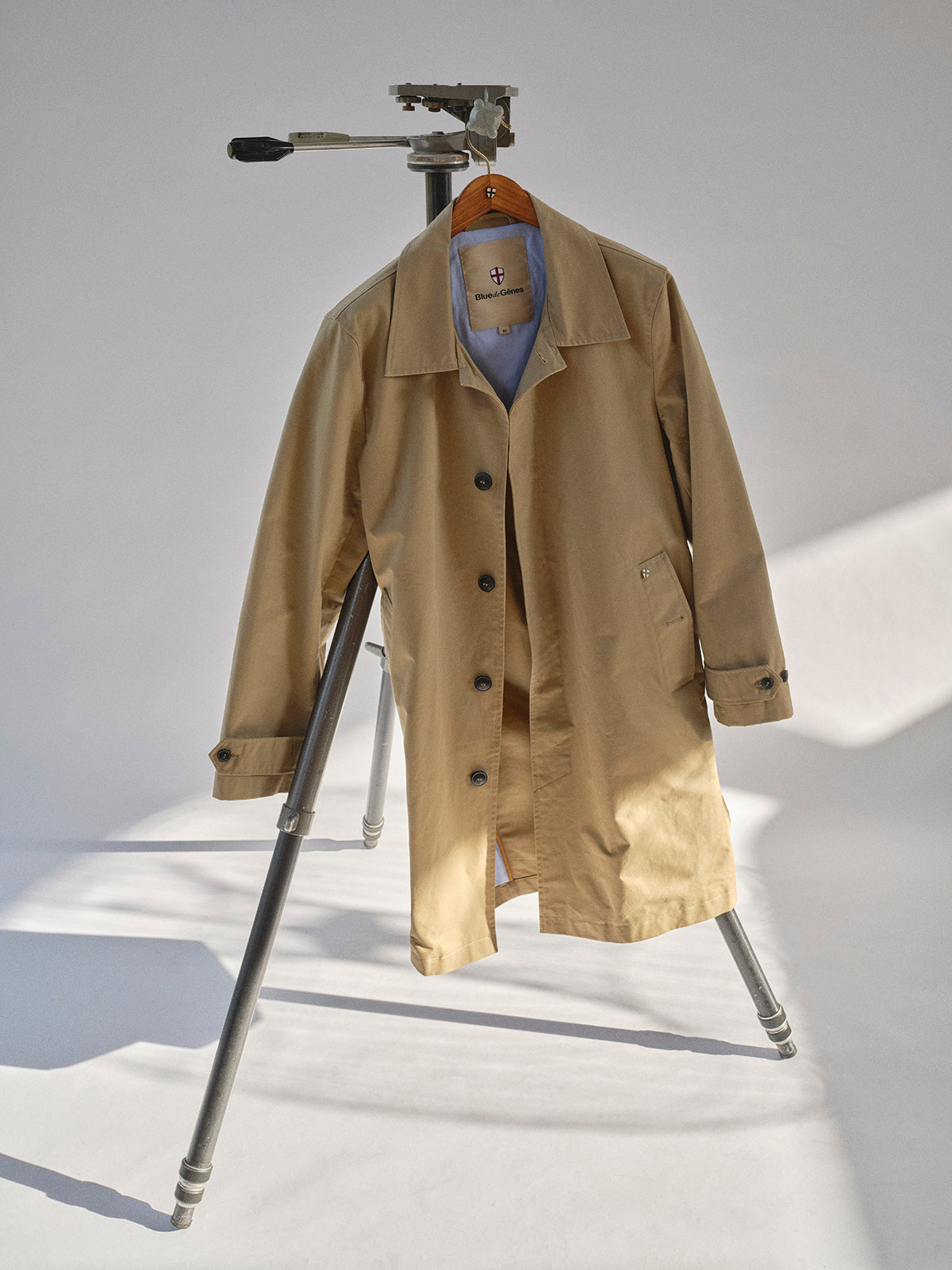 The BGjacques Coat in Pale Khaki by Blue de Gênes, made from heavy cotton, hangs on a tripod stand in a bright room with a simple light background.
