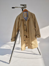 The BGjacques Coat in Pale Khaki by Blue de Gênes, made from heavy cotton, hangs on a tripod stand in a bright room with a simple light background.