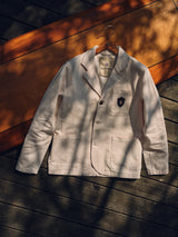 The Blue de Gênes BGbaio Twill Blazer in Chalk, featuring a badge on the chest pocket, hangs on a wooden hanger atop a wood-paneled surface with dappled sunlight and shadows highlighting its classic cream color.