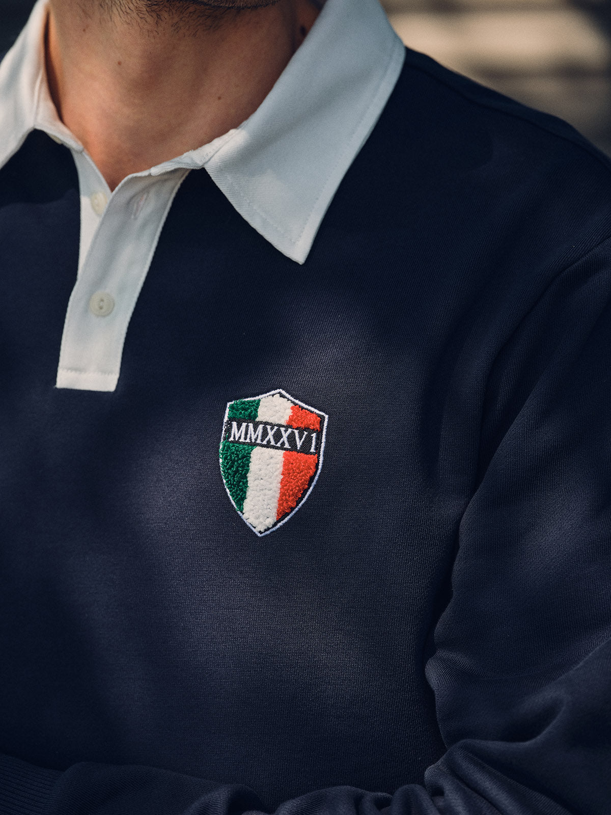 A person wears the Blue de Gênes BGclubhouse Rugby Sweatshirt in navy, from the Clubhouse Champs Collection, featuring a white collar, soft cotton jersey fabric, a chest patch with an Italian flag, and Roman numerals MMXXVI.