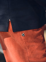 A close-up of a person wearing BGfloyd Cord Shorts - Coral by Blue de Gênes, featuring a small blue cross patch, paired with a navy blue shirt.