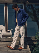 A man stands on wooden steps outside a building with large windows, wearing a blue jacket, white sweater, Blue de Gênes BGpalco Twill Pants in Chalk, and brown shoes; a white chair is nearby.