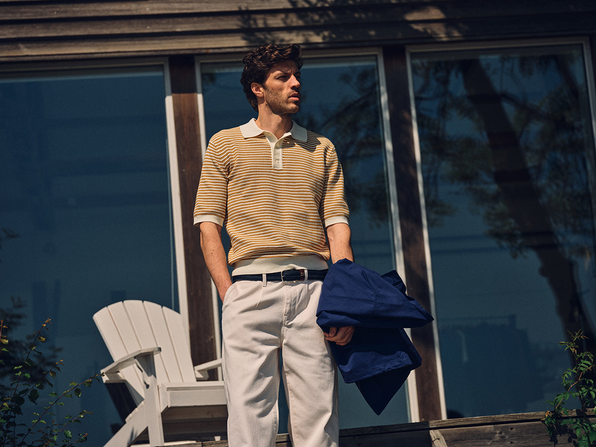 A man stands outside large glass windows wearing the Blue de Gênes BGbob Knit Polo in Sunflower and white pants, holding a blue jacket. A white chair behind him completes his relaxed, nautical look.
