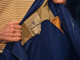 A person holds open a BGcanopy Jacket - Navy Peony by Blue de Gênes, revealing its inner label with the brand’s shield logo, and wears a yellow and white striped top.