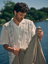 A man stands by a lake, wearing a white embroidered cotton-linen short-sleeve shirt with blue and brown designs. He holds the Blue de Gênes BGjazzy Ola Jacket in Dark Sand, with green trees and blue sky in the background.