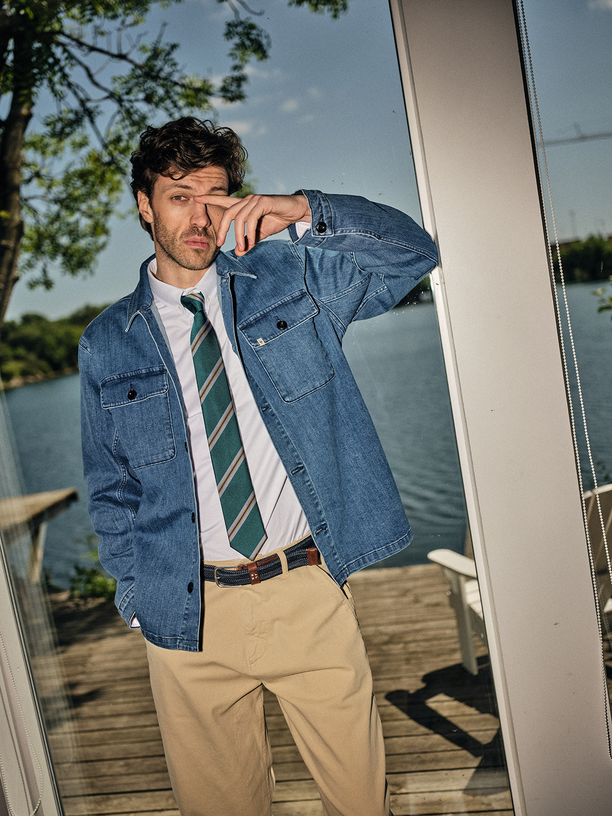 A man stands by a glass door overlooking a lake, wearing the Blue de Gênes BGpontus Denim Overshirt - Used over a white shirt and striped tie with beige pants, one hand in his pocket and the other touching his face.
