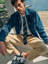A man with tousled hair relaxes by a lake, wearing a Blue de Gênes denim jacket, white shirt, beige pants, green sneakers, and the BGponsy Tie - Green for a stylish men's fashion look.