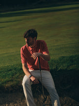 A man in the Blue de Gênes BGwally Diamond Polo - Coral and light pants sits on a sand bunker holding a golf club, with a lush golf course in the background.