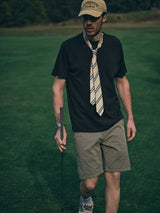 A man in a beige cap, striped tie, black t-shirt, Blue de Gênes BGmenzino Vecchio Shorts in Shadow Green, and sneakers stands on a golf course holding a club. The grassy background is out of focus, highlighting his casual style.