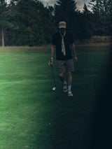 A person walks on a golf course holding a club, wearing Blue de Gênes BGmenzino Vecchio Shorts in Shadow Green along with a cap, dark shirt, and striped tie, surrounded by trees and grass.