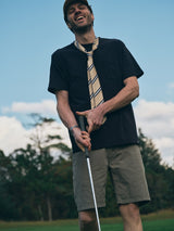 A man in a black T-shirt, striped tie, cap, and Blue de Gênes BGmenzino Vecchio Shorts in Shadow Green laughs while holding a golf club on a lush course with trees and blue sky.