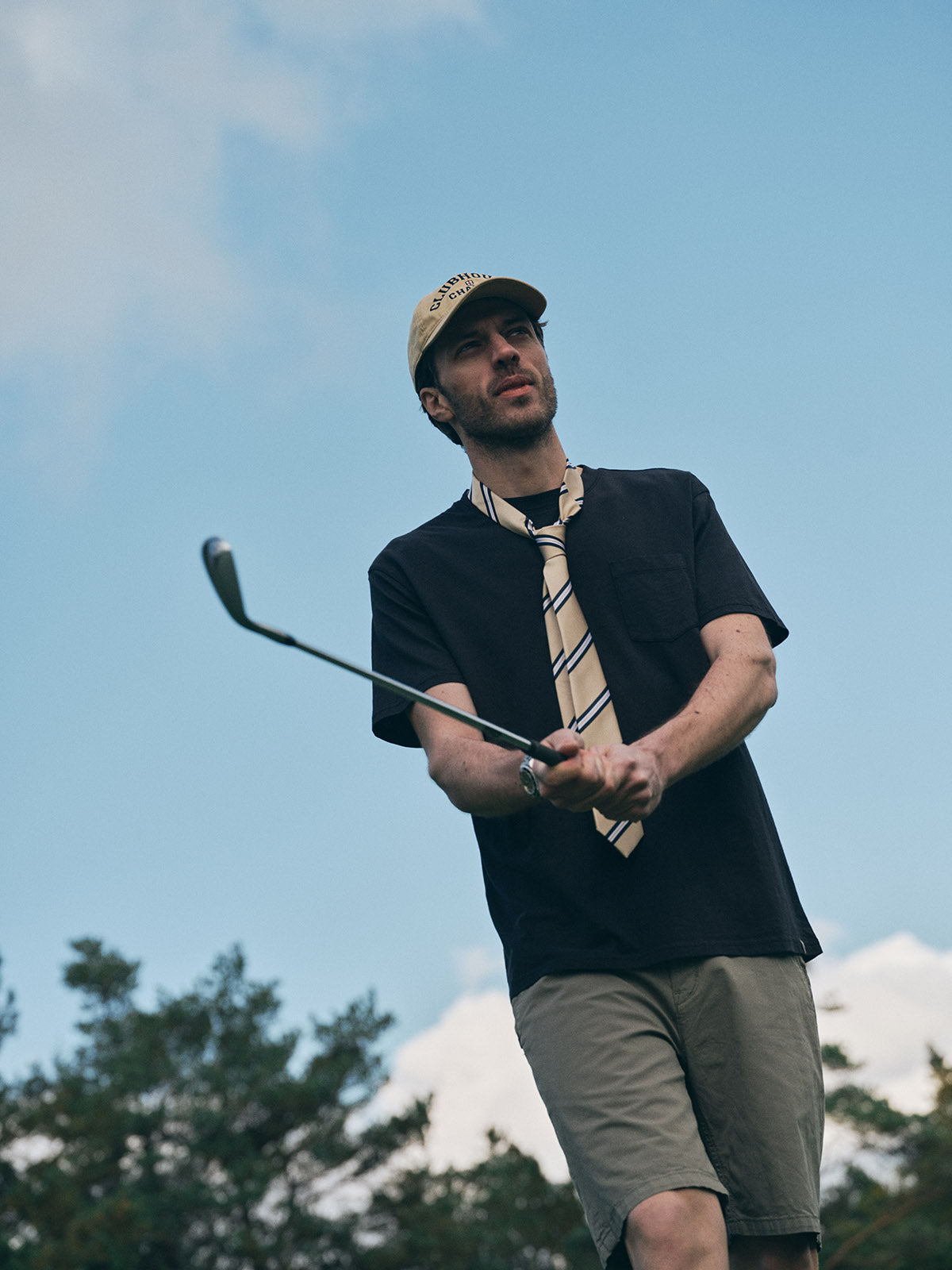 A man in a beige cap, striped tie, khaki shorts, and a Blue de Gênes BGoscar T-Shirt in Jet Black swings a golf club outdoors with trees and a blue sky in the background.