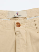 Close-up of BGpalco Rodas Pants in pale khaki by Blue de Gênes, featuring a cotton hemp blend, brown button, fabric label with brand name below a red and white shield logo, visible waistband, belt loops, and relaxed regular fit chinos.