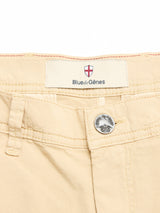 Close-up of BGmenzino Vecchio Shorts in pale khaki by Blue de Gênes, showing the waistband with a label featuring a red and white crest above the brand name, and a silver button engraved with a tree.