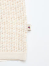 A close-up of the BGwalter S/S Cardigan - Chalk by Blue de Gênes shows cream-colored, textured 100% cotton fabric and a small beige tag with a red and blue shield emblem on the seam against a white background.