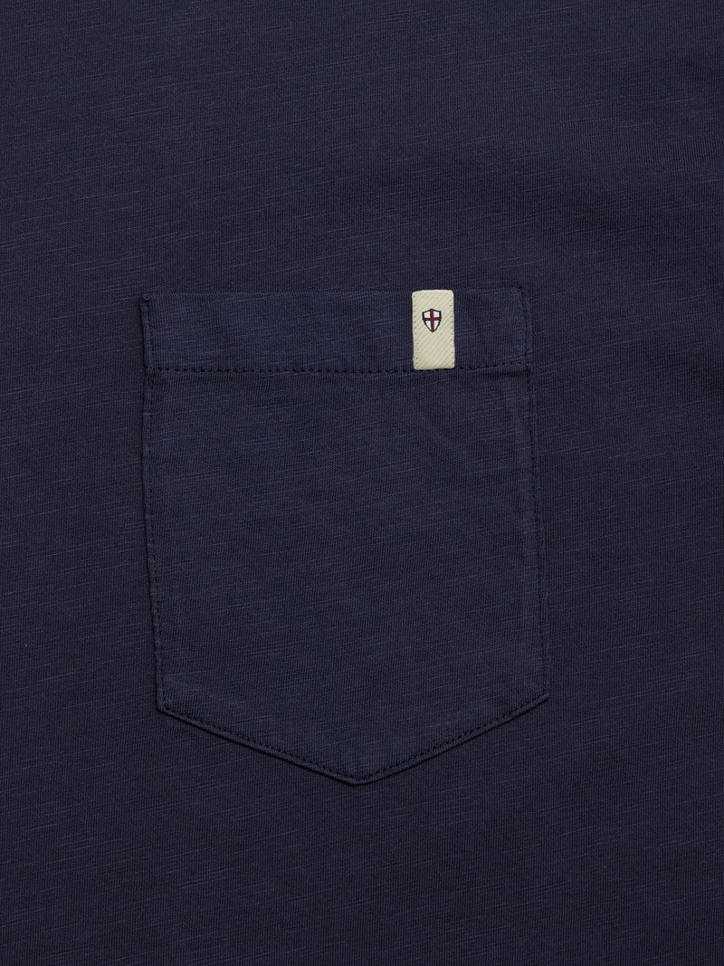 A close-up of the Blue de Gênes BGoscar T-Shirt in dark navy, made from 100% cotton, showing its chest pocket with a cream tag and a red-and-blue shield emblem stitched at the top edge.