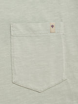 A close-up of the BGoscar T-Shirt - Shadow Green by Blue de Gênes shows its light gray 100% cotton pocket with visible stitching and a small beige tag featuring a red and blue shield emblem on the pocket's upper right corner.