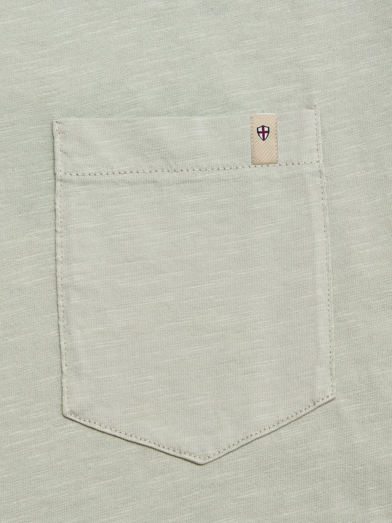 A close-up of the BGoscar T-Shirt - Shadow Green by Blue de Gênes shows its light gray 100% cotton pocket with visible stitching and a small beige tag featuring a red and blue shield emblem on the pocket's upper right corner.