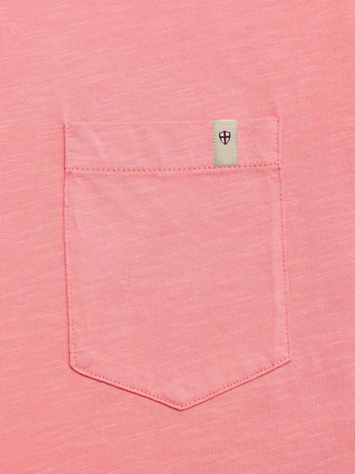 A close-up of the BGoscar T-Shirt in coral by Blue de Gênes, showing the pocket with a small beige tag featuring a black and red shield emblem above it.