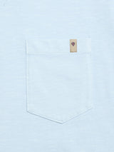 The BGoscar T-Shirt - Blue Fog by Blue de Gênes is a light blue 100% cotton tee with a pocket and a small beige tag at the top right corner, featuring a red and black shield emblem.