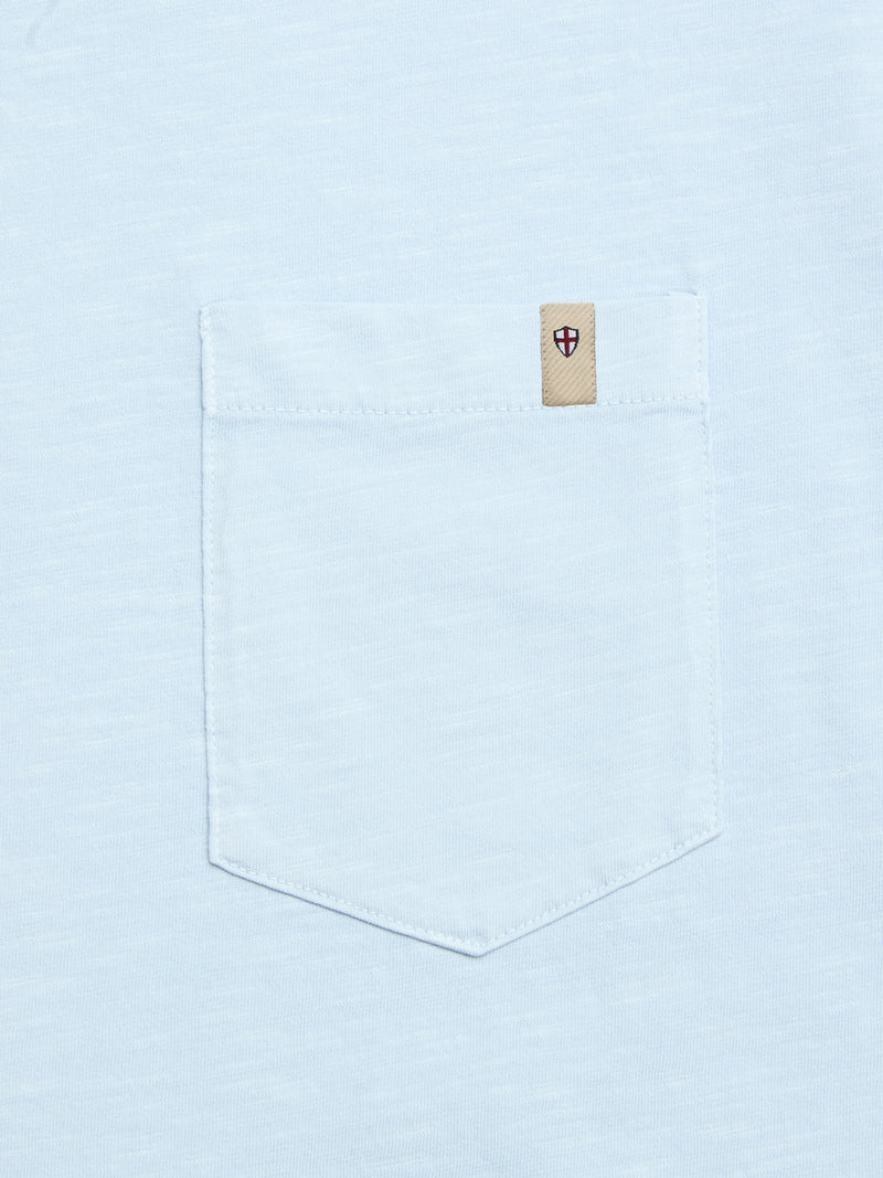 The BGoscar T-Shirt - Blue Fog by Blue de Gênes is a light blue 100% cotton tee with a pocket and a small beige tag at the top right corner, featuring a red and black shield emblem.