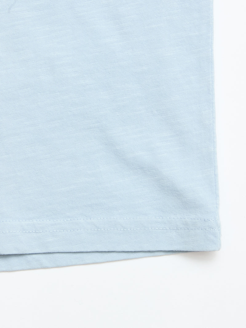 Close-up of the bottom hem of the Blue de Gênes BGoscar T-Shirt - Blue Fog, a 100% cotton crewneck, showcasing stitching and fabric texture against a white background.