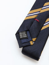 A close-up of the Blue de Gênes BGponsy Tie - Dark Navy, a silk tie with diagonal yellow and white stripes, shows its pointed end and a tag stating 100% silk in English, French, and German.