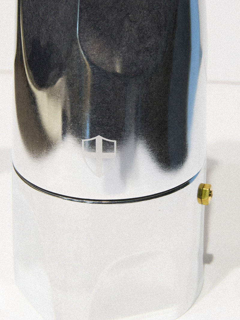 A close-up of the BDG Coffee Maker - Grey by Blue de Gênes, featuring a shiny metallic finish, reflective surface, small shield emblem, and brass-colored bolt set against a white background.
