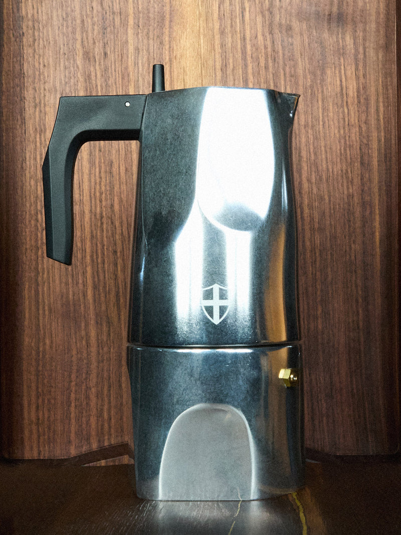 The BDG Coffee Maker - Grey by Blue de Gênes, a modern stainless steel espresso pot with a black handle and minimalist shield logo, sits on a dark wooden surface against a matching background, reflecting light.