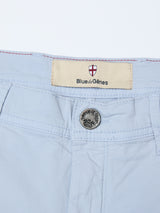 Close-up of BGmenzino Vecchio Shorts in Blue Fog, featuring a light blue waistband, metallic button, and a "Blue de Gênes" label topped by a red and white shield emblem.