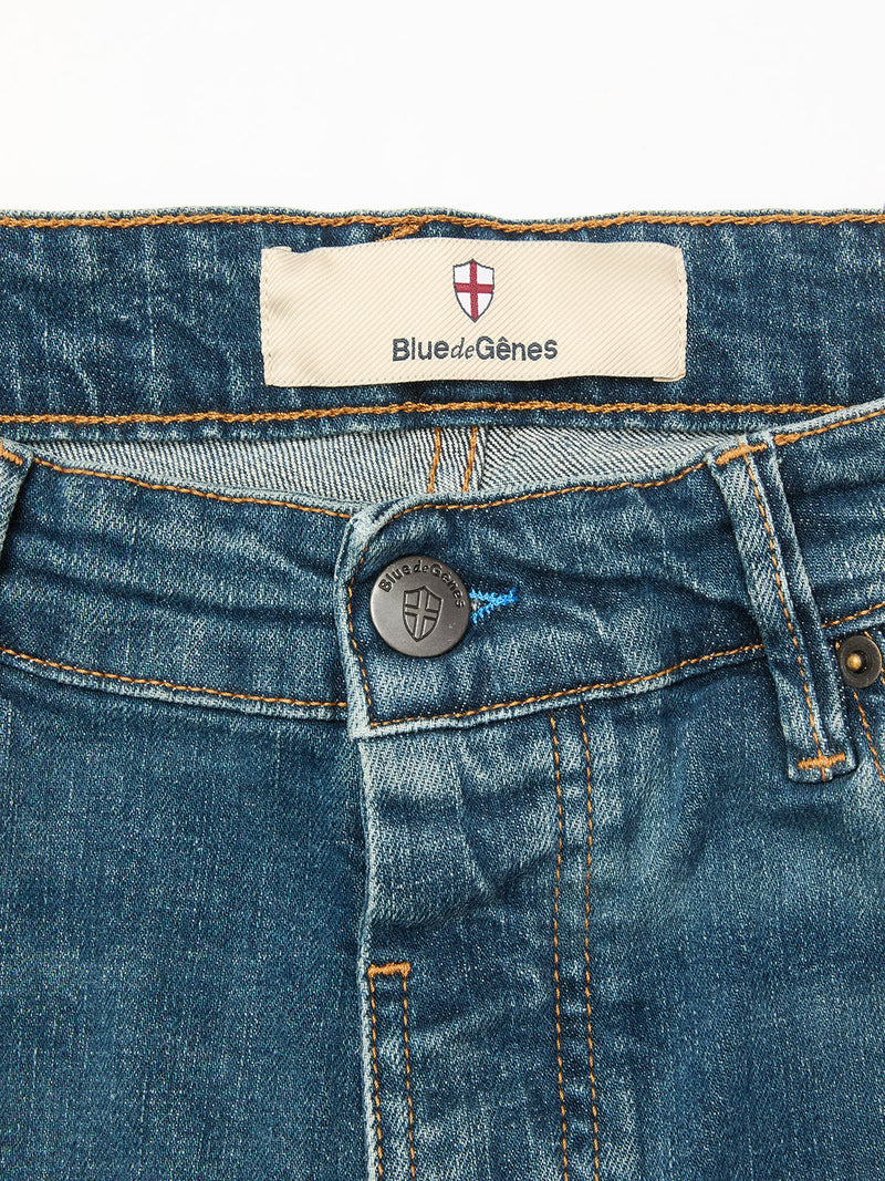 Close-up of Blue de Gênes Vinci Topaz Vintage Jeans - Used, highlighting the blue denim waistband, black shield-embossed button, orange stitching, and a white Blue de Gênes label with red and white shield logo.