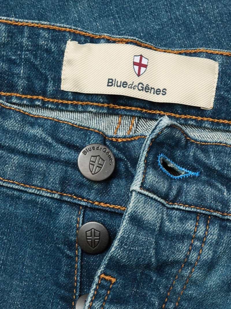 Close-up of Vinci Topaz Vintage Jeans - Used by Blue de Gênes in blue comfort stretch denim, featuring exposed branded buttons, a label with a red cross above the brand name, orange stitching, and a blue buttonhole.