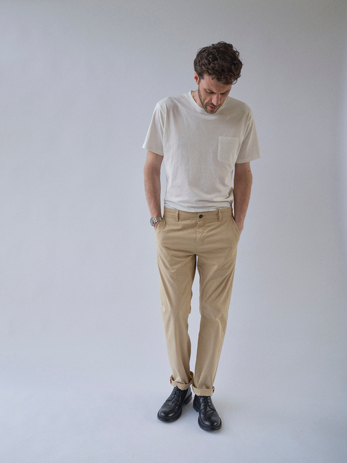 A man stands against a light background, looking down with his hands in the pockets of his Blue de Gênes BGmenzo Vecchio Chino - Pale Khaki, paired with a white T-shirt, black shoes, and a watch.
