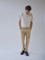 A man stands against a light background, looking down with his hands in the pockets of his Blue de Gênes BGmenzo Vecchio Chino - Pale Khaki, paired with a white T-shirt, black shoes, and a watch.