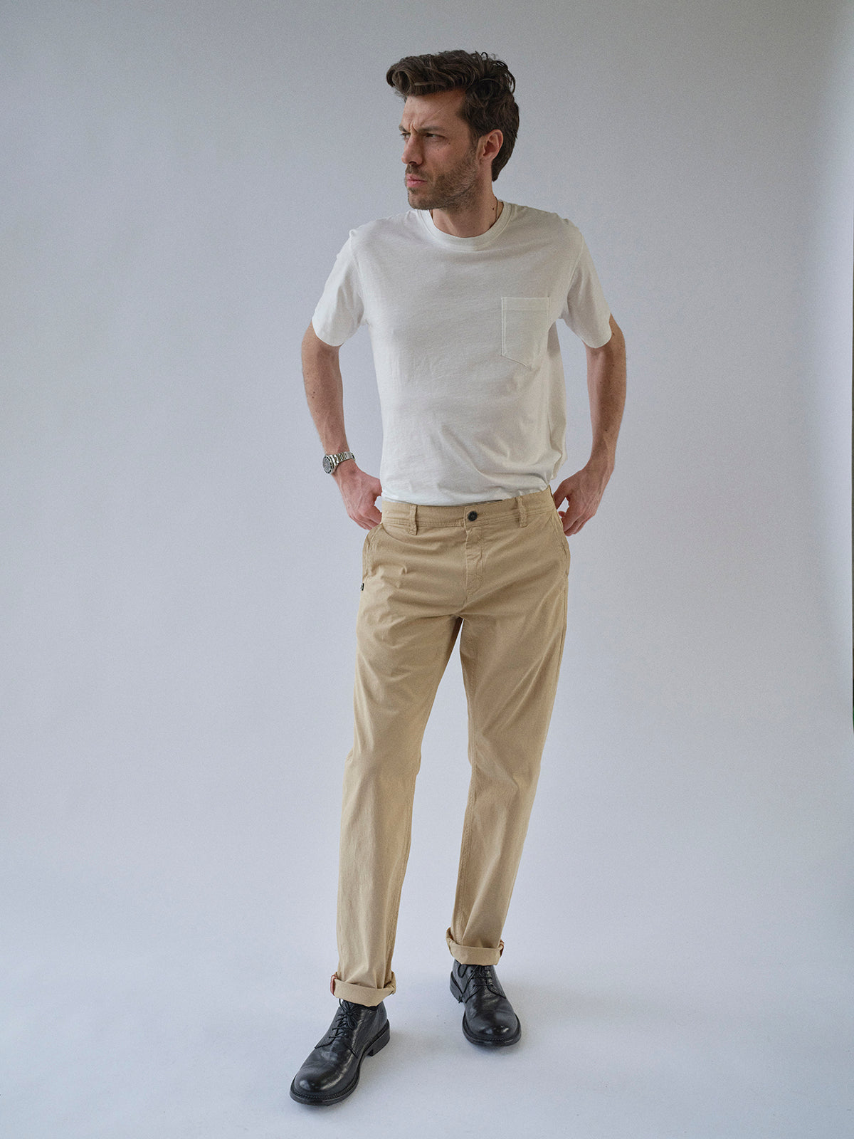 A man with short hair stands against a plain light background wearing the Blue de Gênes BGmenzo Vecchio Chino in Pale Khaki, a white t-shirt, black shoes, watch on his left wrist, hands in pockets, and looking slightly to the side.