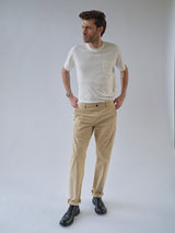 A man with short hair stands against a plain light background wearing the Blue de Gênes BGmenzo Vecchio Chino in Pale Khaki, a white t-shirt, black shoes, watch on his left wrist, hands in pockets, and looking slightly to the side.