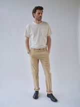 A man stands against a light background wearing a white T-shirt, black shoes, and Blue de Gênes BGmenzo Vecchio Chino in Pale Khaki, with his hands in his pockets and looking slightly to the side.