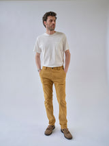 A man stands against a plain light background wearing a white t-shirt, brown shoes, and Blue de Gênes BGpaulo Pavia Pant - Sunflower, with his hands in his pockets and looking slightly to his left.