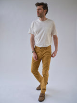 A man stands against a plain background wearing a white short-sleeve tee, Blue de Gênes BGpaulo Pavia Pant in Sunflower, and brown shoes, with one hand in his pocket and looking off to the side.