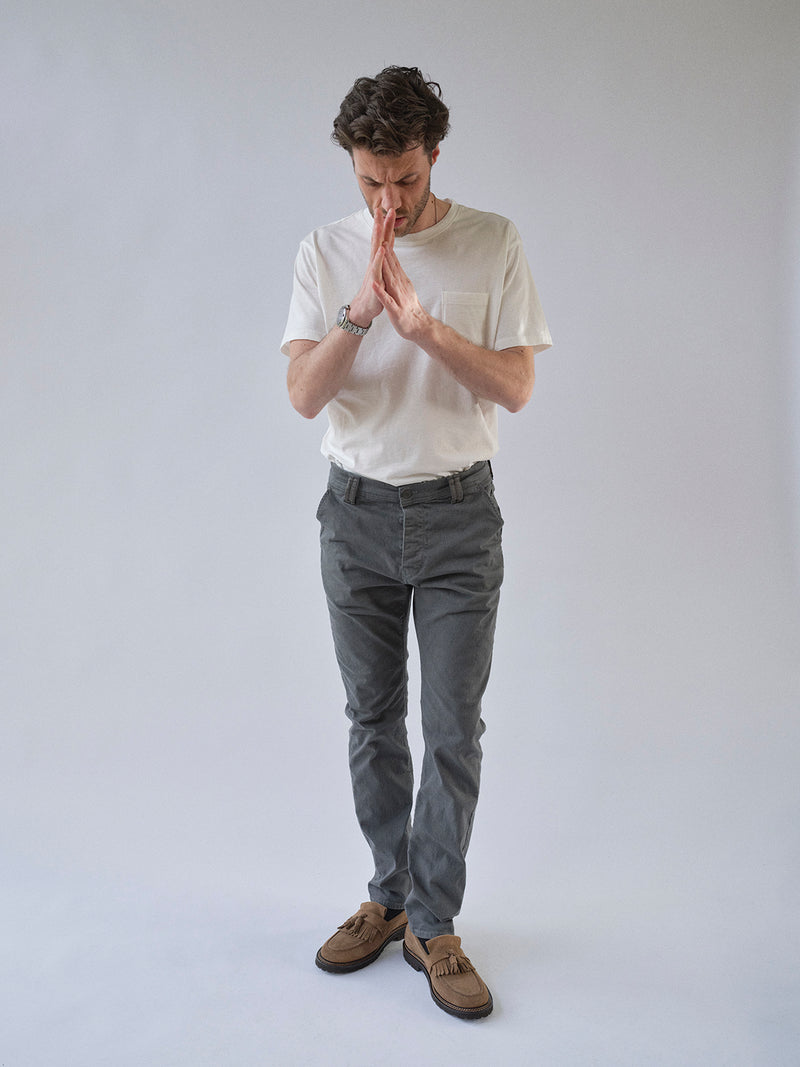 A man in a white t-shirt, BGpaulo Pavia 1 V2 Chino - Charcoal by Blue de Gênes, and brown shoes stands against a plain light background, looking down with his hands pressed together in front of his face as if deep in thought.