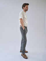 A man stands in profile against a plain background, wearing a white t-shirt, Blue de Gênes BGpaulo Pavia 1 V2 Chino in charcoal, and brown shoes, with his hands in his pockets.