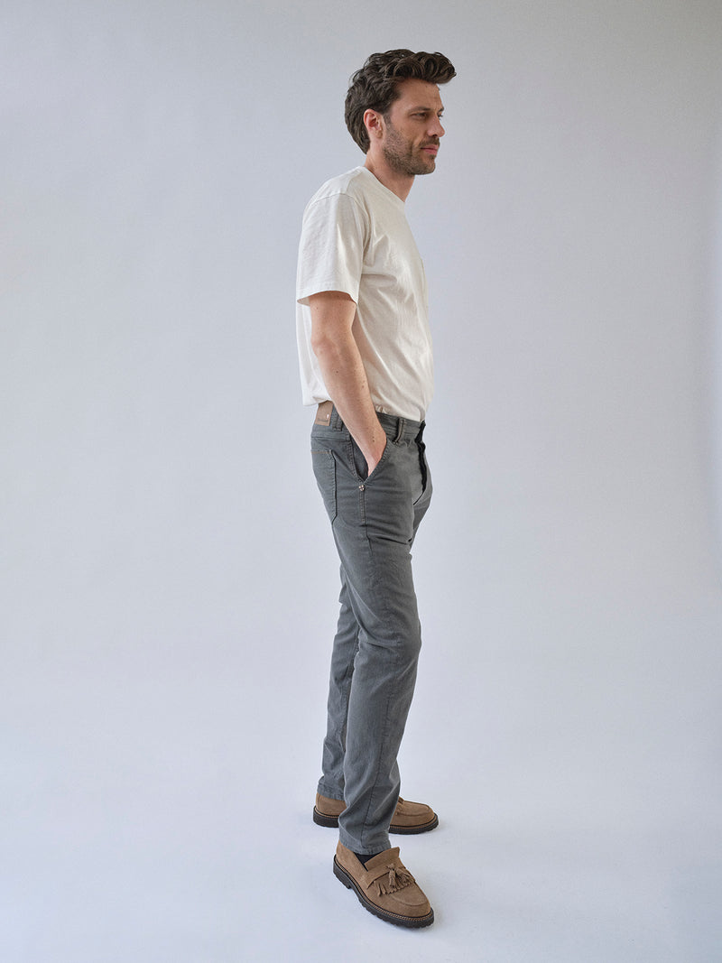 A man stands in profile against a plain background, wearing a white t-shirt, Blue de Gênes BGpaulo Pavia 1 V2 Chino in charcoal, and brown shoes, with his hands in his pockets.