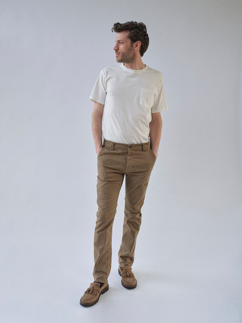 A man stands against a plain light background wearing a white t-shirt, Blue de Gênes BGpaulo Pavia 1 V2 Chino - Fudge pants, and brown loafers. His hands are in his pockets as he looks left with a relaxed posture.