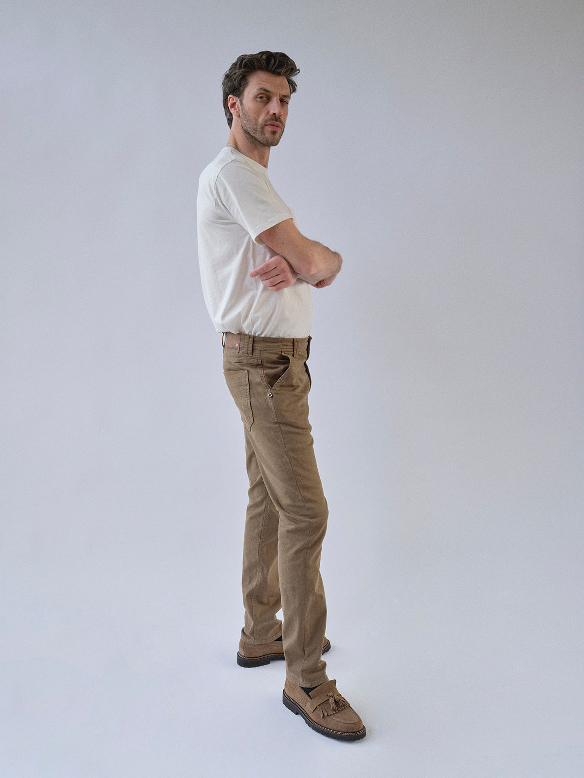 A man stands sideways with arms folded, wearing a white t-shirt and Blue de Gênes BGpaulo Pavia 1 V2 Chino - Fudge pants with brown shoes, against a plain light gray background.