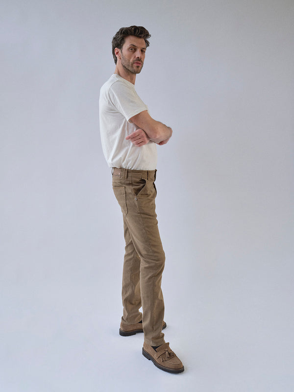 A man stands sideways with arms folded, wearing a white t-shirt and Blue de Gênes BGpaulo Pavia 1 V2 Chino - Fudge pants with brown shoes, against a plain light gray background.